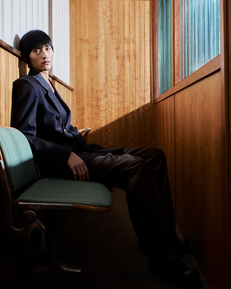 Portrait of Asian man in blue suit sitting in vintage upholstered chair indoors with wood paneled walls and stained-glass window.