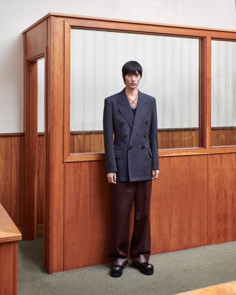 Asian male model poses in a double-breasted blazer, wide-leg pants, and black shoes against a wood-paneled wall with a glass partition.