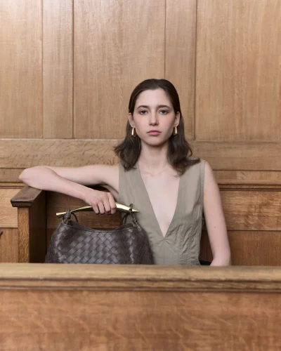 Model wearing a V-neck top posing next to a woven leather handbag against a wood-paneled wall
