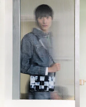 Model Minsoo Lee viewed through a fluted glass door wearing a grey jacket and a black and white woven crossbody bag.