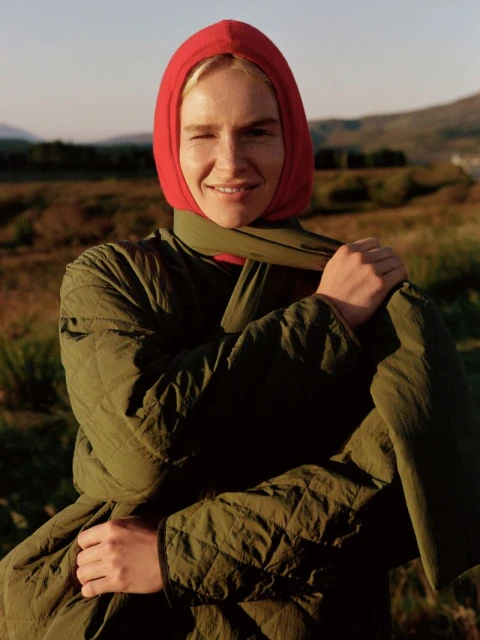 Woman in a quilted jacket and red headscarf smiles in a medium outdoor shot against a background of a field and distant hills.
