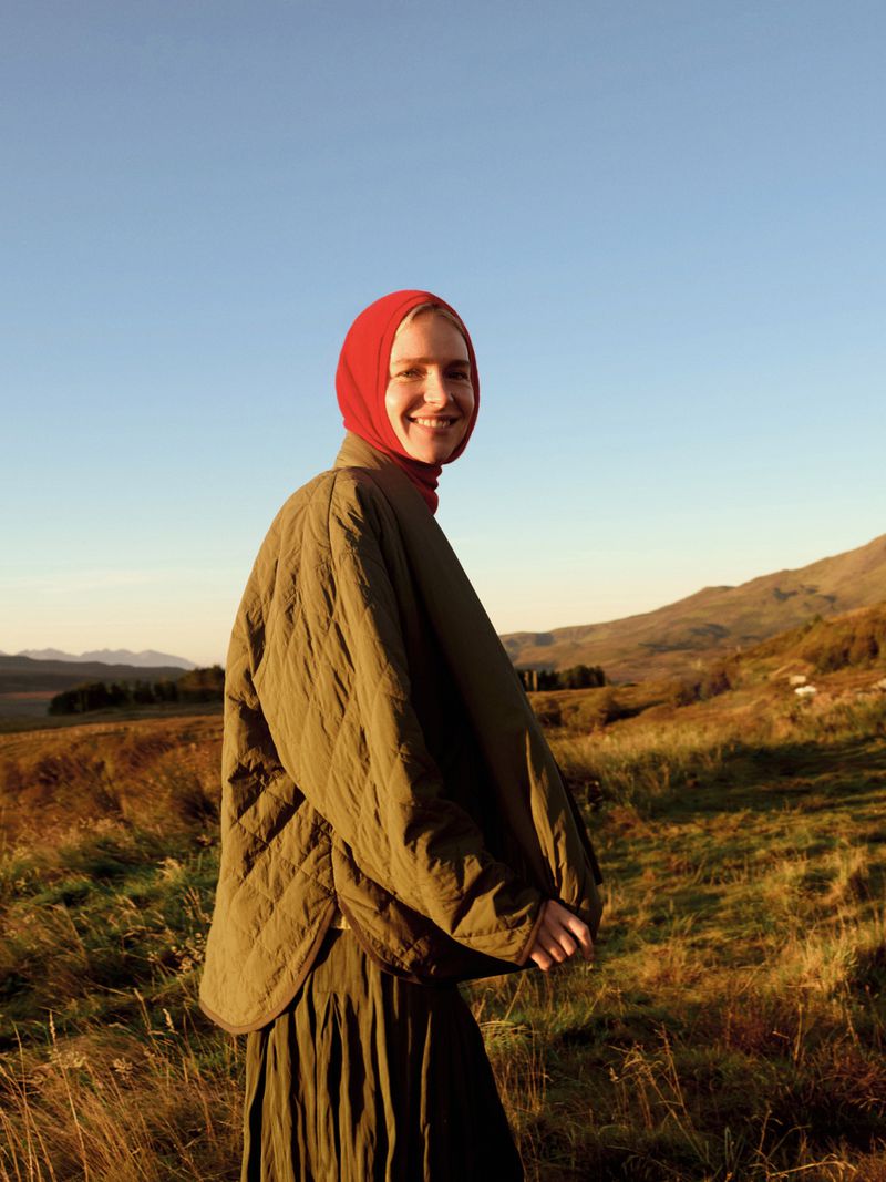 Outdoor portrait of smiling woman in olive green quilted jacket and red hood, set against a backdrop of grassy hills and clear blue sky.