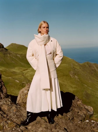 Model in a cream puffer jacket, matching scarf, and skirt standing on a rocky mountain slope overlooking the sea.