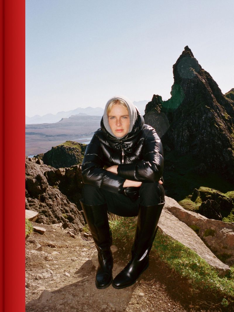 Model wearing a black glossy puffer jacket and tall boots sitting outdoors on a rocky mountain slope