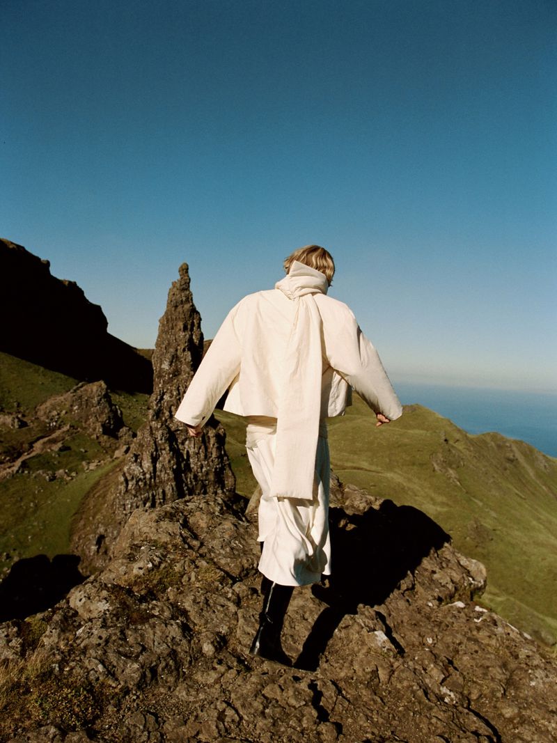 Fashion shot of a person in white clothing walking on a rocky mountain ridge on a sunny day with blue sky and green landscape background.