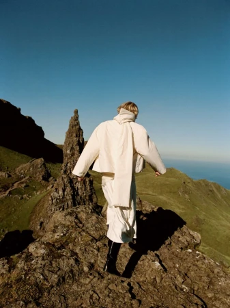Fashion shot of a person in white clothing walking on a rocky mountain ridge on a sunny day with blue sky and green landscape background.