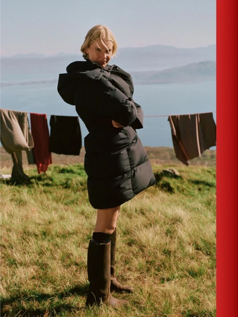 A fair-skinned woman in a large black puffer coat and boots stands in a grassy field, clothes on a line, and a mountain and lake backdrop.