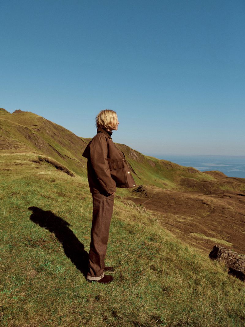 Adult stands on a grassy hillside wearing a brown jacket and pants, overlooking rolling green hills and a blue sky in natural light.
