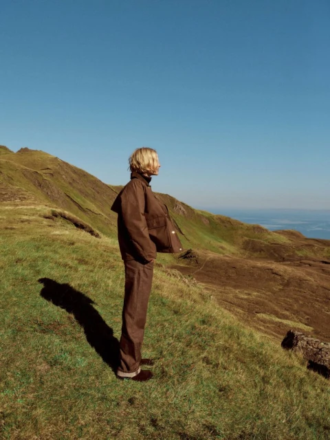 Adult stands on a grassy hillside wearing a brown jacket and pants, overlooking rolling green hills and a blue sky in natural light.