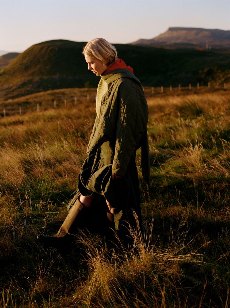 A woman walks through a golden field wearing a quilted coat, with rolling green hills and a mountain visible in the background.