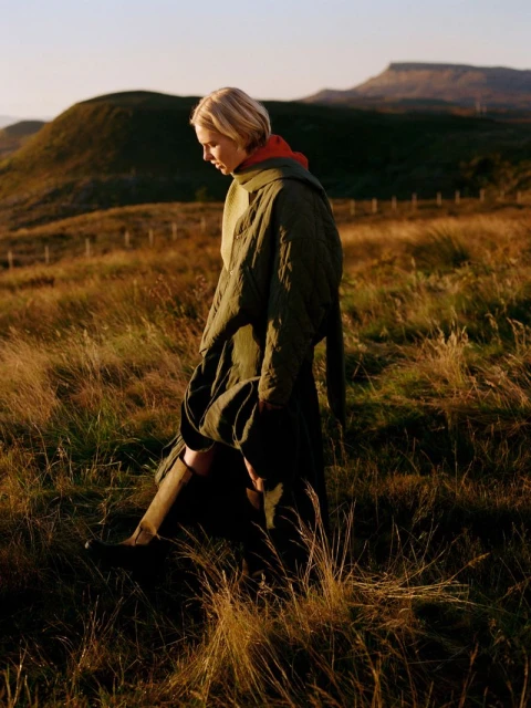 A woman walks through a golden field wearing a quilted coat, with rolling green hills and a mountain visible in the background.