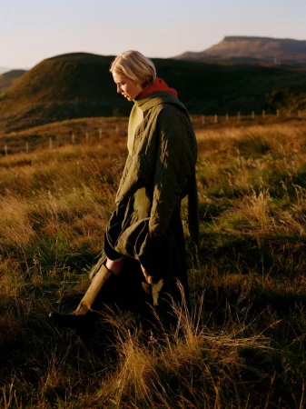 A woman walks through a golden field wearing a quilted coat, with rolling green hills and a mountain visible in the background.