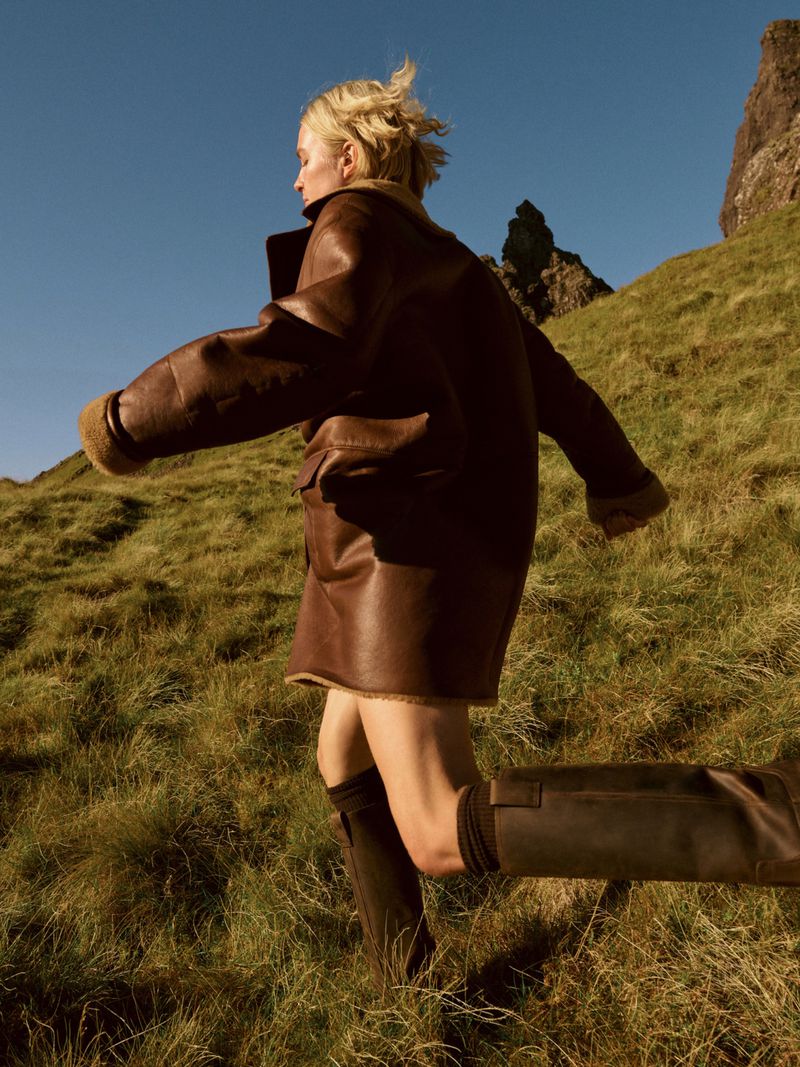 A woman runs down a grassy hill in a brown coat and black boots, with a rocky outcrop and blue sky in the background, outdoors.