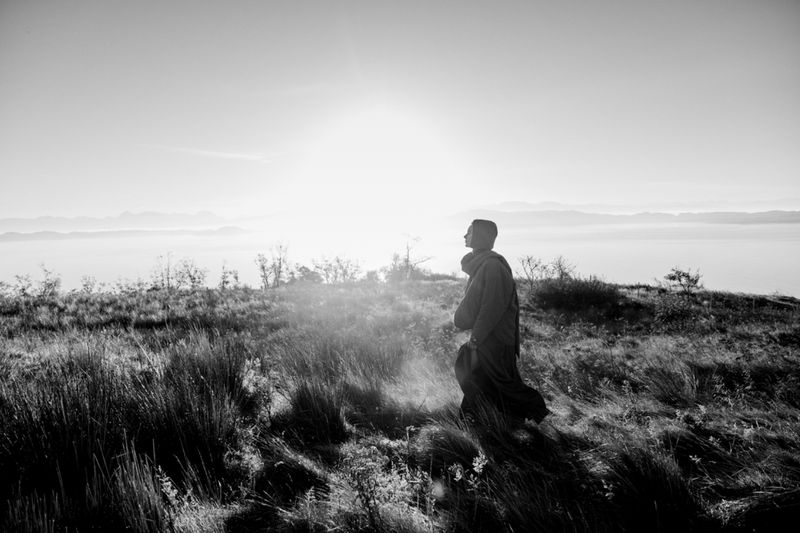 Monochrome image of a monk in robe standing in a grassy field with mountain backdrop, bathed in sunlight, evoking serenity.