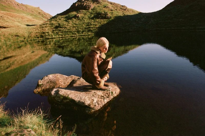 Young woman in aviator jacket sits on rock in a serene lake surrounded by grassy hills in a tranquil outdoor setting with natural lighting.