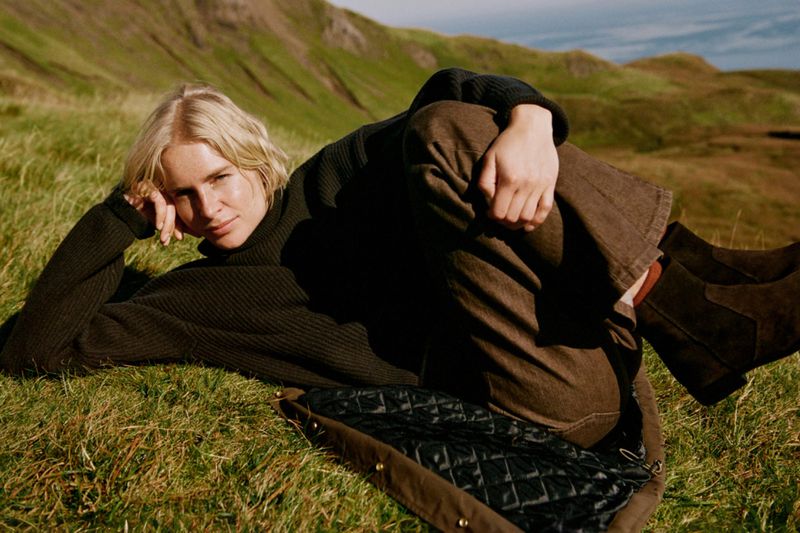 Blonde woman in a green sweater and brown pants rests on a grassy hill in a serene outdoor setting with mountains in the background.