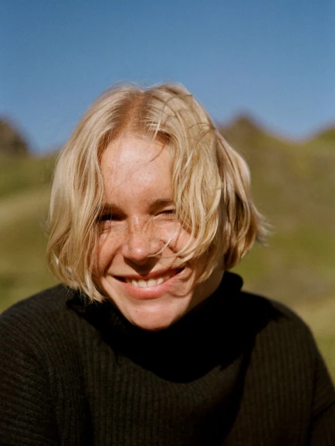 Blond woman with freckles smiles outdoors against a background of a grassy hill and blue sky on a sunny day in a portrait shot.