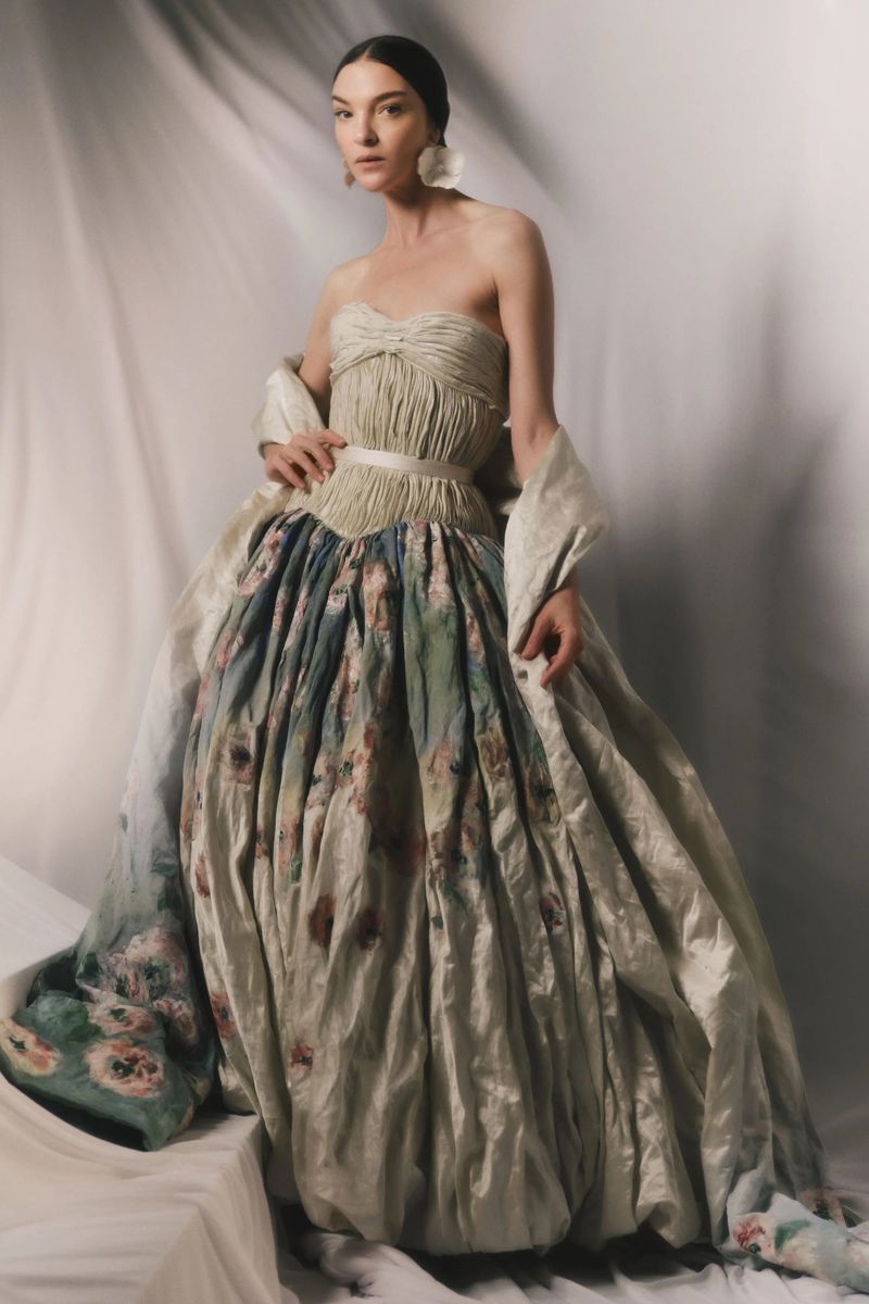 Model Mariacarla Boscono wearing a voluminous strapless floral evening gown against a draped fabric backdrop.