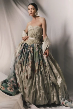 Model Mariacarla Boscono wearing a voluminous strapless floral evening gown against a draped fabric backdrop.