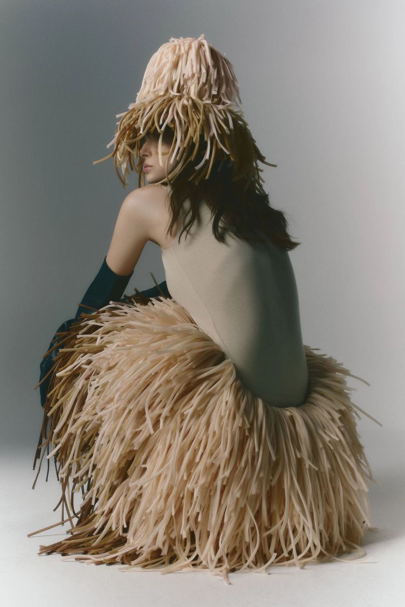 Model wearing beige bodysuit with fringed skirt and matching hat, in studio setting, posing against gray background.