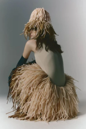 Model wearing beige bodysuit with fringed skirt and matching hat, in studio setting, posing against gray background.