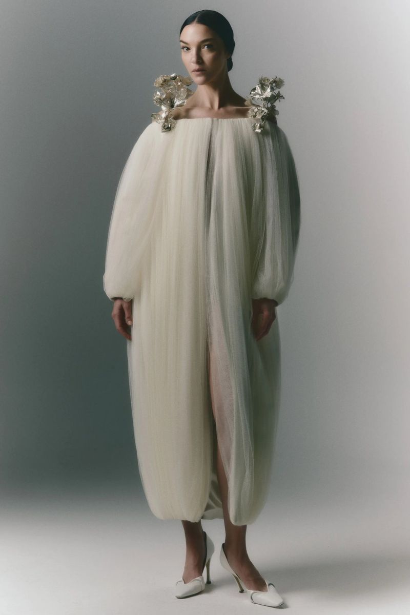 A woman in a sculptural, ivory gown with floral shoulder embellishments poses in a full-length studio shot against a neutral backdrop.