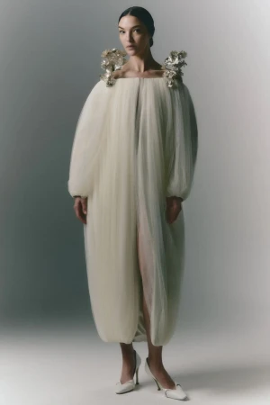A woman in a sculptural, ivory gown with floral shoulder embellishments poses in a full-length studio shot against a neutral backdrop.