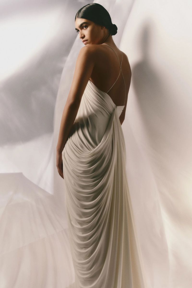 Woman wearing a flowing white gown, shot from behind in a bright indoor setting with draped fabric, showcasing elegant fashion and design.