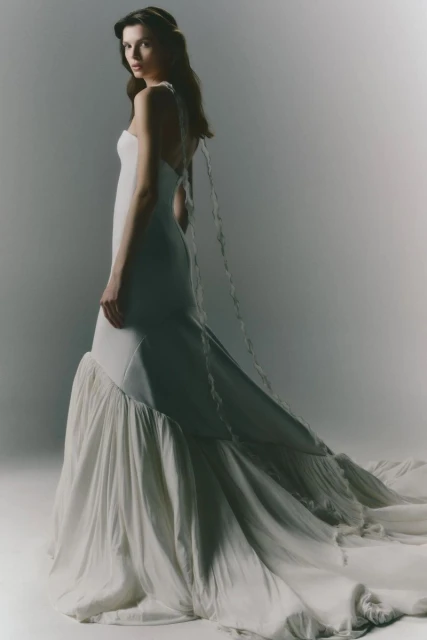 Brunette woman in flowing white wedding dress with cascading train and decorative ribbon straps posing in a bright studio setting.