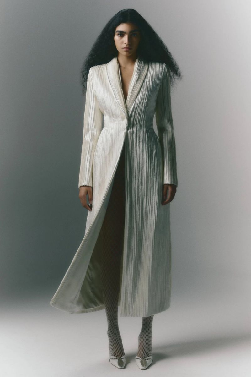 Full-length portrait of an adult female fashion model wearing a long, textured white coat, fishnet stockings, and white heels in a studio.