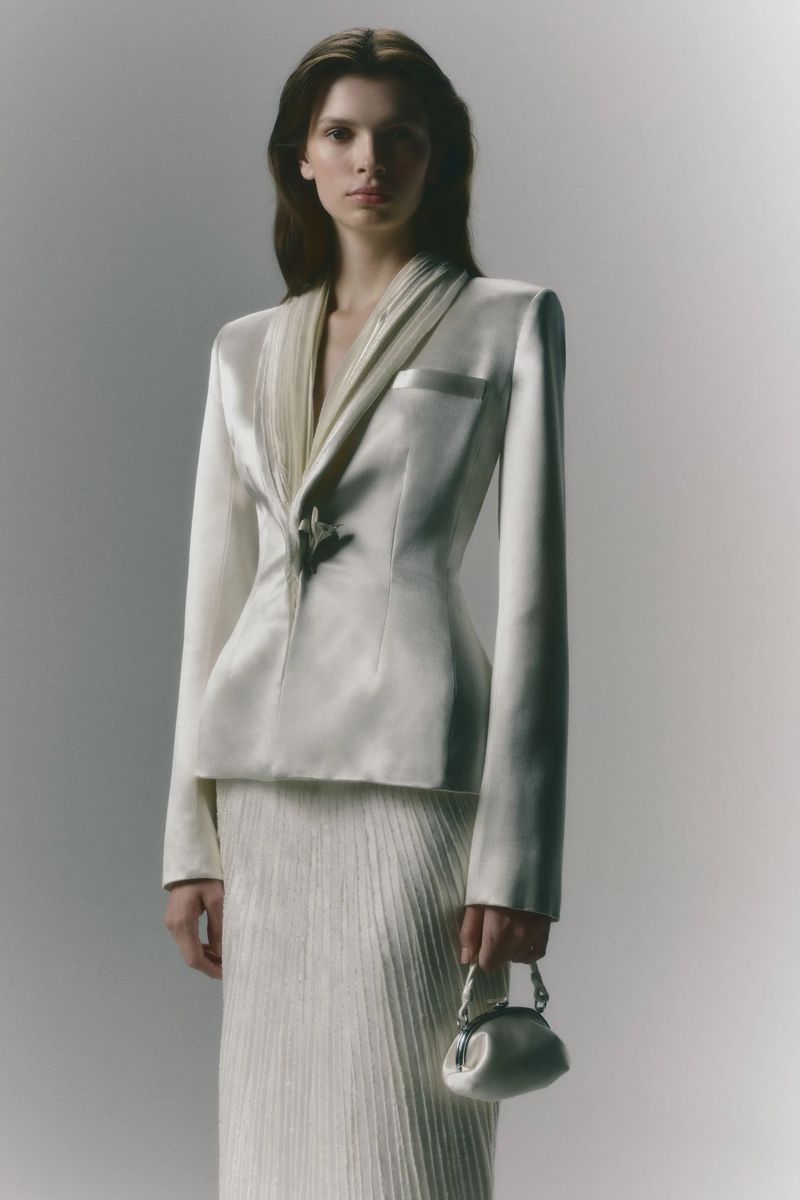 A poised fashion model in an off-white suit with a satin finish, complemented by a small handbag, poses against a bright studio backdrop.