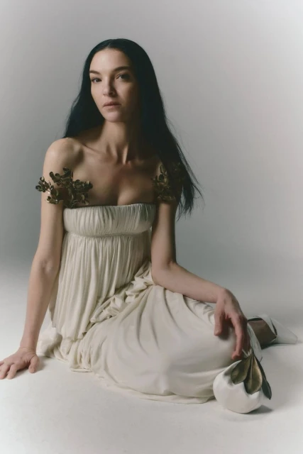 Elegant woman in off-white dress with floral accents sitting on the floor in a photo studio with soft, diffused lighting.