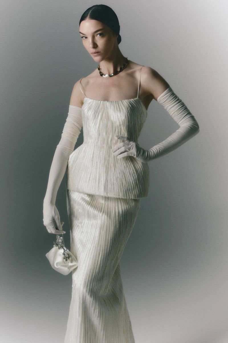 An elegant woman poses in a cream-colored, floor-length evening gown with matching gloves and handbag against a neutral backdrop.