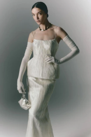An elegant woman poses in a cream-colored, floor-length evening gown with matching gloves and handbag against a neutral backdrop.