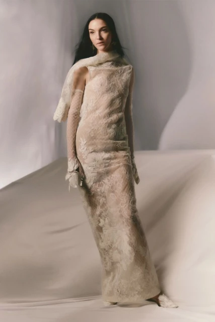 A woman poses in a floor-length lace dress, wearing a lace scarf and gloves, against a light background in a fashion studio setting.