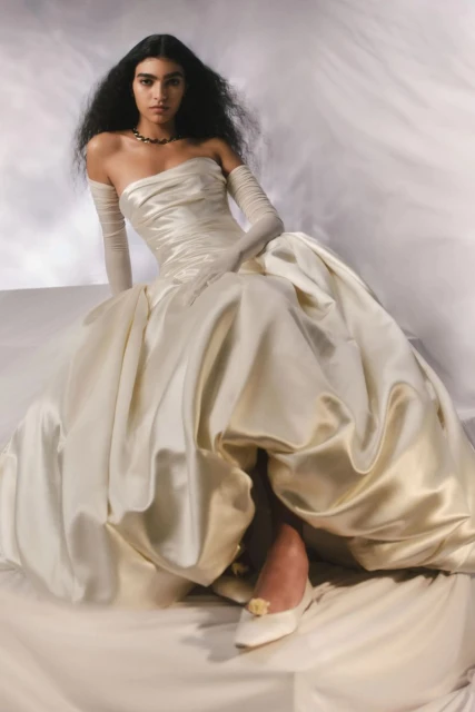 Elegant woman with long dark hair poses in a strapless ivory gown and gloves against a soft backdrop in a studio fashion shot.