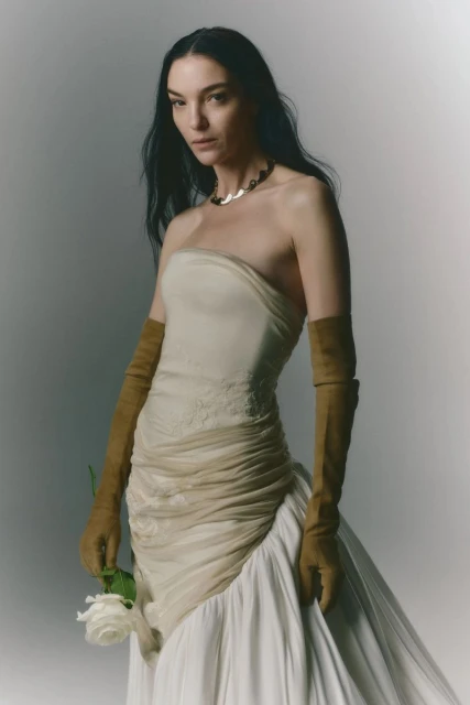 Elegant woman in strapless dress and gloves poses with a white rose in a light studio setting, showcasing fashion and beauty.
