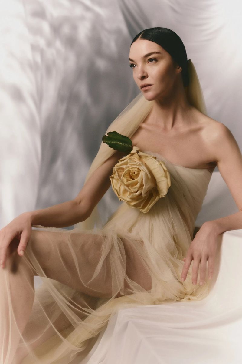 An adult woman sits in a draped, sheer gown, with dark hair pulled back. The gown has a rose embellishment on the shoulder.