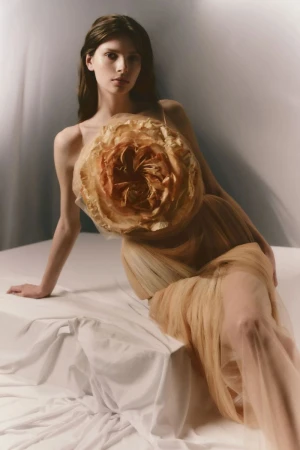 Model reclining in a nude tulle dress with a large sculptural floral rosetta applique against a soft studio backdrop.