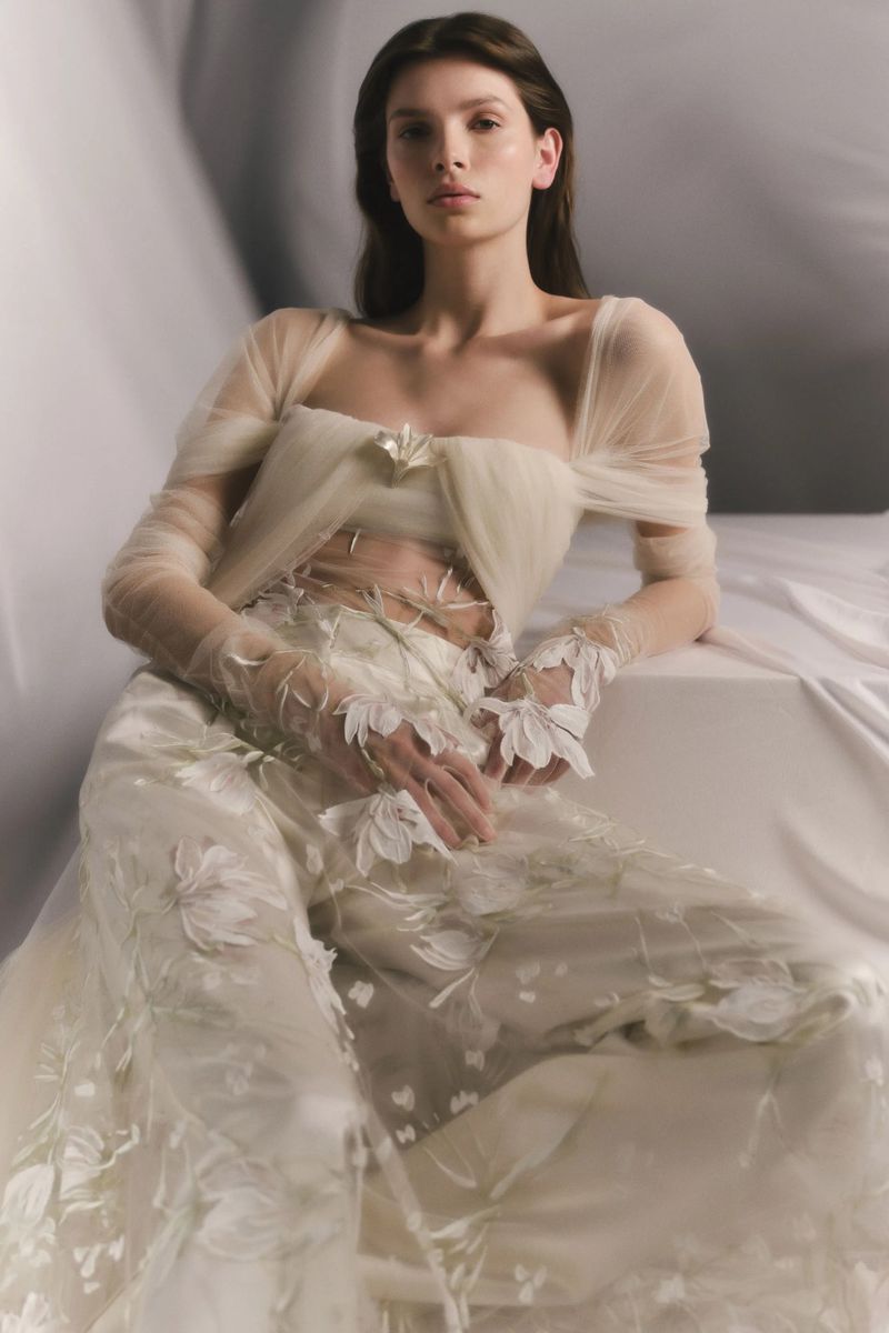 Model Anita Pozzo seated in a sheer off-the-shoulder cream tulle top with embroidered trousers against a draped fabric backdrop.