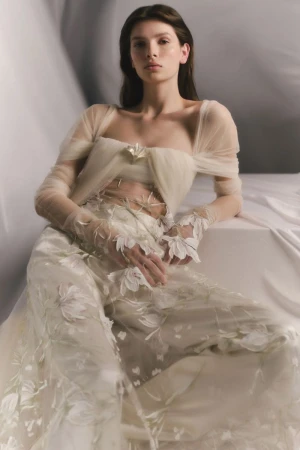 Model Anita Pozzo seated in a sheer off-the-shoulder cream tulle top with embroidered trousers against a draped fabric backdrop.