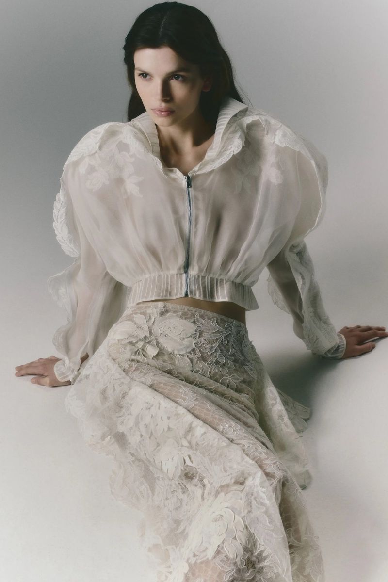 Model Anita Pozzo seated wearing a sheer cropped white jacket and matching embroidered lace skirt on a white studio backdrop.