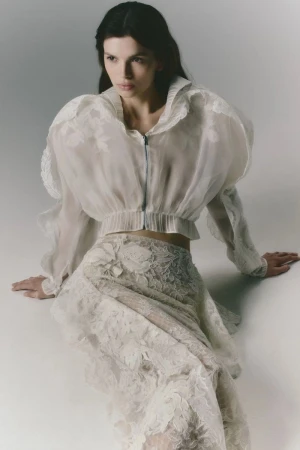 Model Anita Pozzo seated wearing a sheer cropped white jacket and matching embroidered lace skirt on a white studio backdrop.
