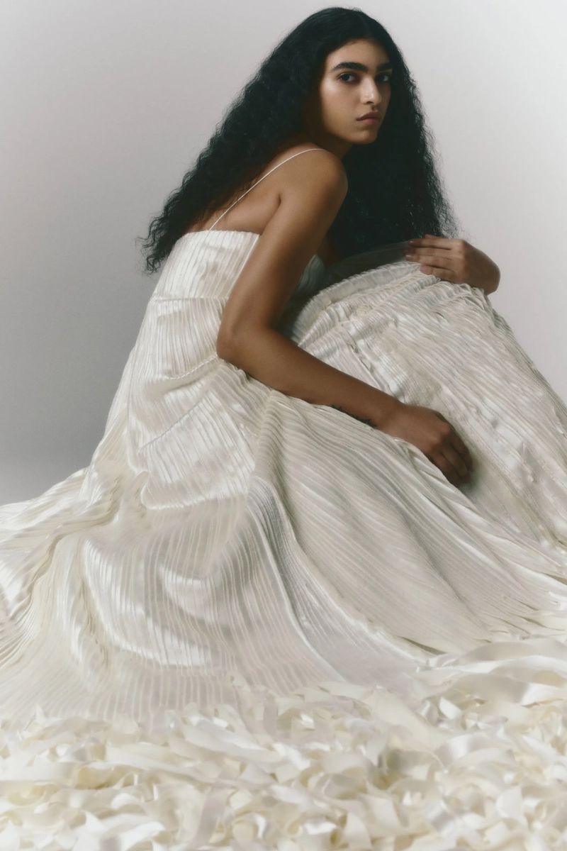 A woman with dark hair is sitting, posing in a formal white dress. The dress has texture and a flowing train on a white background.