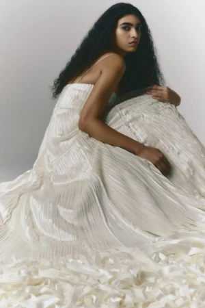 A woman with dark hair is sitting, posing in a formal white dress. The dress has texture and a flowing train on a white background.