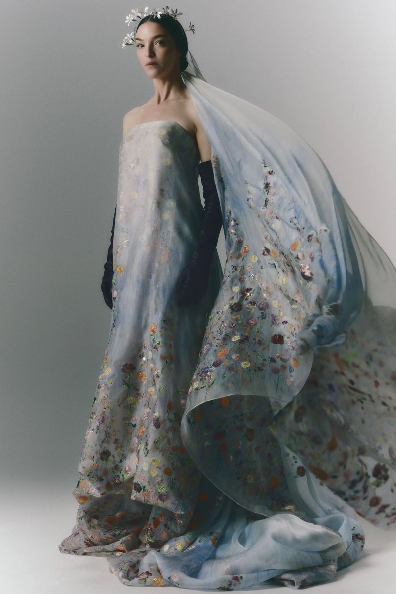 Elegant woman in ethereal gown with floral accents and a flowing veil, captured in a full-length studio shot against a bright backdrop.