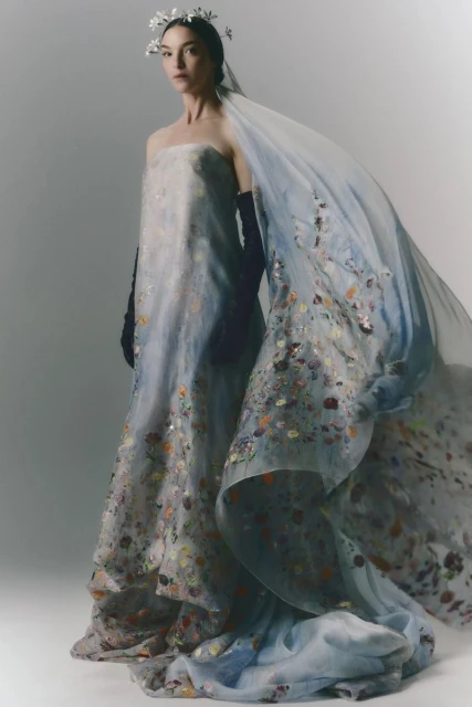 Elegant woman in ethereal gown with floral accents and a flowing veil, captured in a full-length studio shot against a bright backdrop.