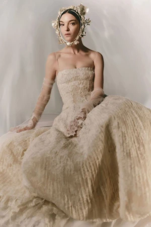 Model wearing a lace dress and floral headpiece seated against a neutral draped background for Danielle Frankel FW24.