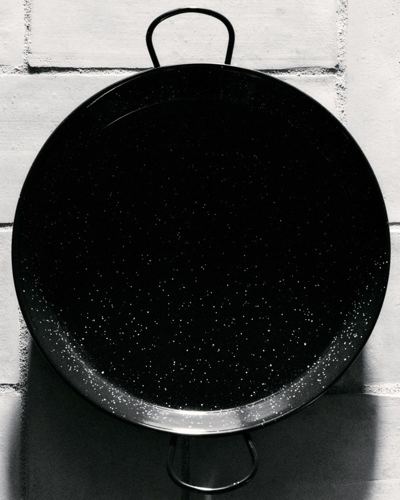 Monochrome photo of a black paella pan with handles on a brick wall, featuring a shiny surface and textured background.