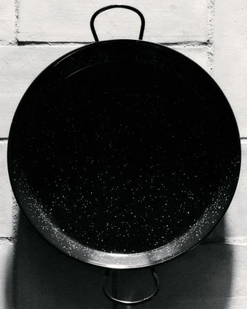 Monochrome photo of a black paella pan with handles on a brick wall, featuring a shiny surface and textured background.